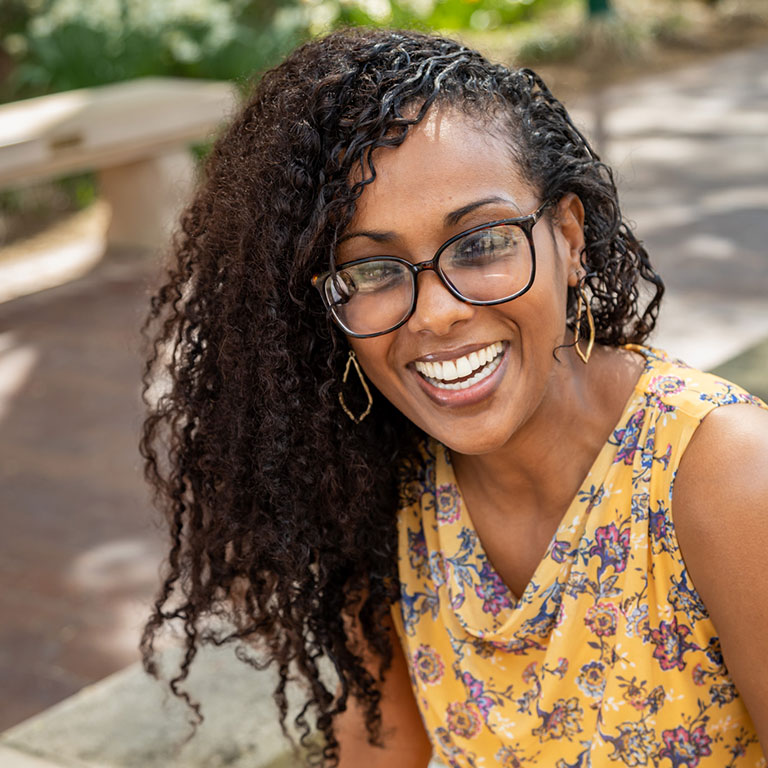 A headshot of Muna Adem, who wears a yellow blouse and poses outside.