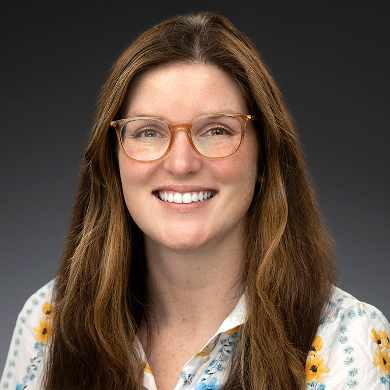 a headshot of Maeve Bartiss, who wears a flower-patterned shirt and poses against a dark background.