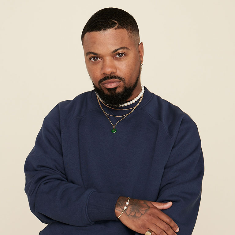 A headshot of Cedric Hudson, who wears a blue sweatshirt and poses against a beige backdrop.