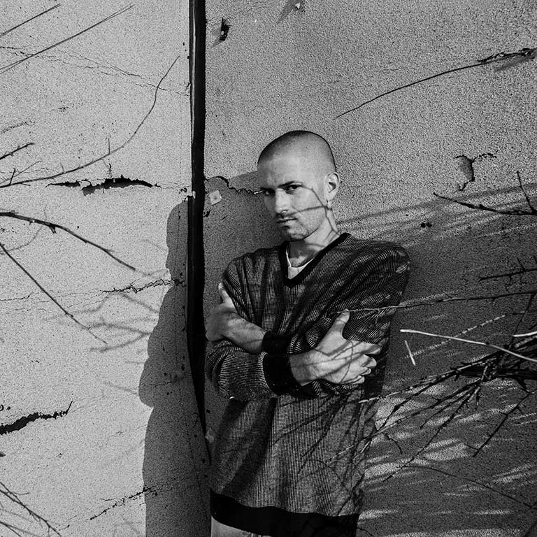 A photo of Andrés Mario de Varona, which is black and white. De Varona wears a gray sweater and poses against a cracked wall.