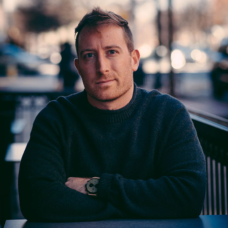 Brian Fitzgerald sits at a table outdoors in a city, wearing a dark blue sweater and a wristwatch.