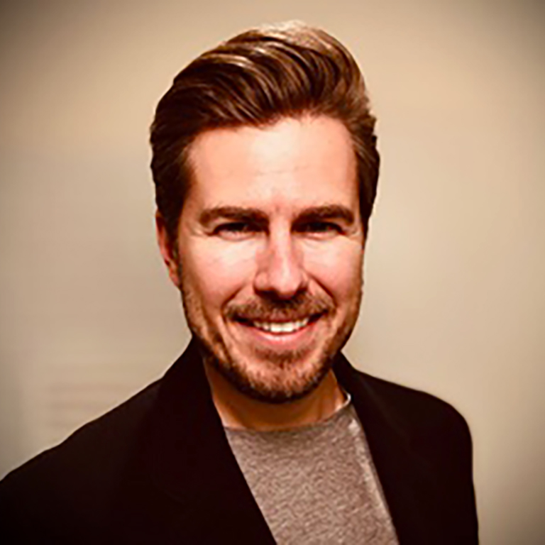 A headshot of Miles Taylor, who wears a black blazer and poses against a beige background.